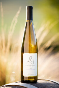 A bottle of Chasselas Doré sitting on a wine barrel in the golden hour with ornamental grasses and the Estate vineyard in the background.