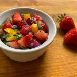 Strawberry salsa, including strawberries, red onion, yellow bell pepper, cilantro and jalapeño, finely diced and mixed together in a dish with whole strawberries next to it on a cutting board