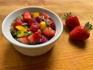 Strawberry salsa, including strawberries, red onion, yellow bell pepper, cilantro and jalapeño, finely diced and mixed together in a dish with whole strawberries next to it on a cutting board