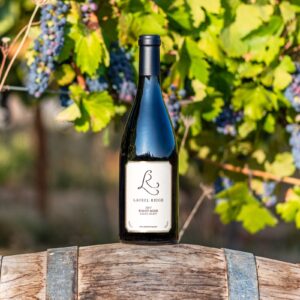 a bottle of Laurel Ridge pinot noir sitting on a wine barrel in front of some vines in the Estate vineyard.